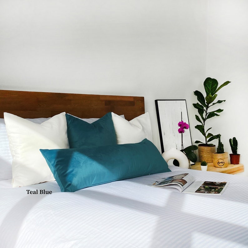 May include: Teal blue velvet lumbar pillow on a white bed with white pillows and a wooden headboard. The bed has a white and blue striped bedspread. There is a magazine on the bed and a potted plant on a side table.