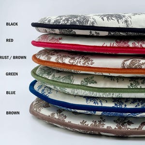 May include: A stack of round cushions with a toile pattern in shades of gray and brown. Each cushion has a different colored border: black, red, rust/brown, green, blue, and brown. The cushions are stacked on top of each other.
