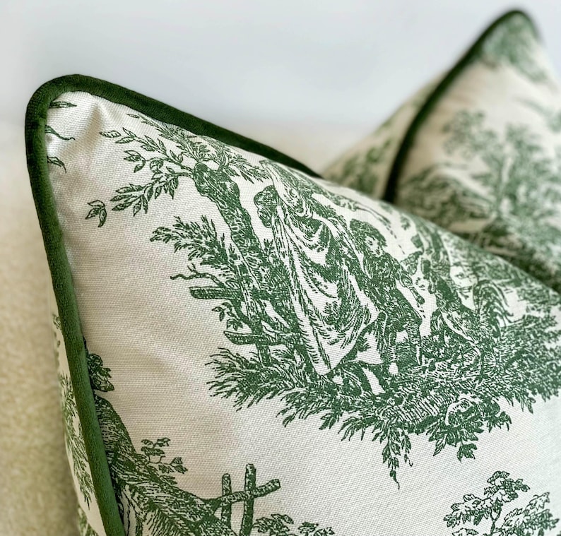 May include: Two decorative throw pillows with a green and white toile pattern. The pillows feature a detailed pastoral scene and are trimmed with a dark green velvet border. The pillows are a light cream color.