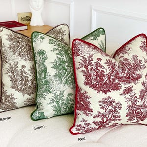 May include: Three decorative throw pillows in brown, green, and red. Each pillow features a cream-colored background with a detailed toile pattern depicting figures and trees. The pillows have contrasting colored piping and are arranged on a white sofa.