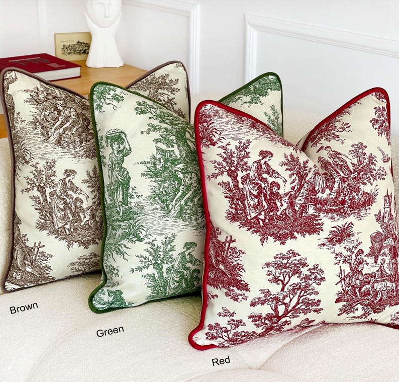 May include: Three decorative throw pillows in brown, green, and red. Each pillow features a cream-colored background with a detailed toile pattern depicting figures and trees. The pillows have contrasting colored piping and are arranged on a white sofa.