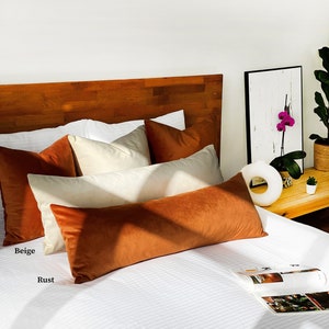 May include: A white bed with a wooden headboard and a rust colored velvet bolster pillow. There are also several beige and white pillows on the bed.