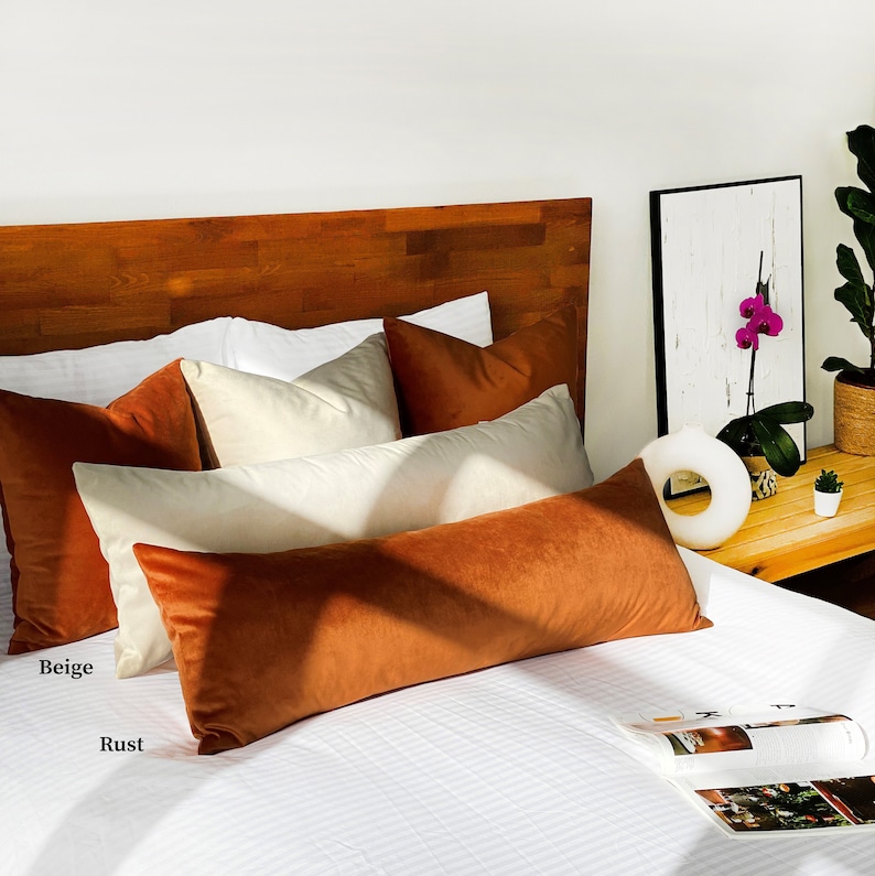May include: A white bed with a wooden headboard and a rust colored velvet bolster pillow. There are also several beige and white pillows on the bed.