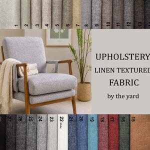 Peut inclure: Tissu d'ameublement texturé aspect lin vendu au mètre, présenté avec un fauteuil gris clair aux accoudoirs en bois et un plaid crème. L'image inclut une palette de couleurs numérotée de 1 à 28, montrant divers échantillons de tissus dans des tons neutres, bleus, rouges, verts et marron.