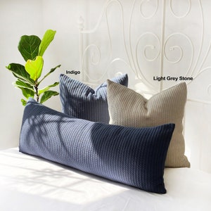May include: Three decorative pillows on a white bed. The pillows are made of a textured fabric and are in the colors indigo, light grey stone, and white. The pillows are arranged in a row, with the indigo pillow in the back, the light grey stone pillow in the middle, and the white pillow in the front.