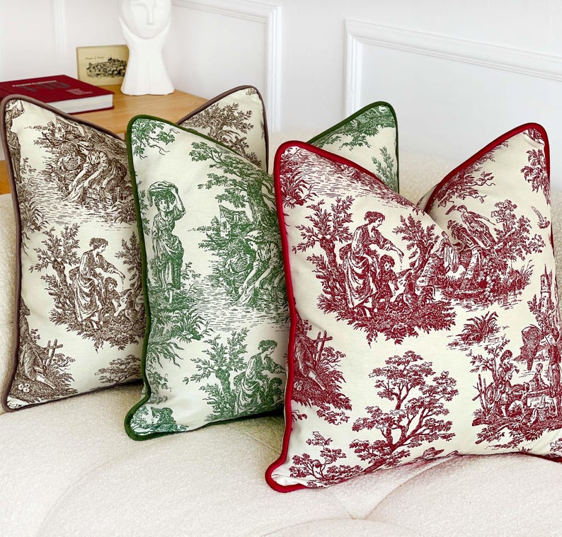 K&ouml;nnte beinhalten: Vier dekorative Kissen mit Toile-de-Jouy-Muster. Die Kissen haben einen cremefarbenen Hintergrund mit Szenen in Braun, Gr&uuml;n und Rot. Jedes Kissen hat eine kontrastierende Bord&uuml;re in Braun, Gr&uuml;n bzw. Rot. Die Kissen sind auf einem wei&szlig;en Sofa angeordnet.