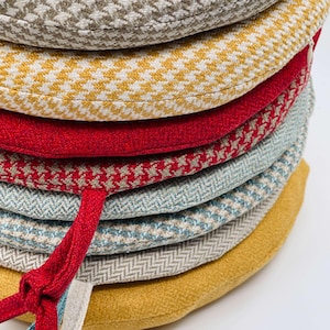May include: A stack of round seat cushions in various colors and patterns. The cushions include houndstooth, herringbone, and solid designs in shades of yellow, red, blue, and beige. A red tie is attached to one cushion.