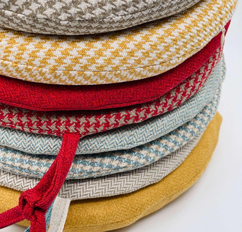 Puede incluir: Una pila de cojines redondos para asientos en varios colores y patrones. Los cojines incluyen telas de pata de gallo, espiga y lisas en tonos de amarillo, rojo, azul y beige. Una cinta roja est&aacute; unida a un coj&iacute;n.