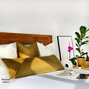 May include: A mustard yellow velvet lumbar pillow on a bed with white bedding and a wooden headboard. The pillow is placed on top of two white pillows. There is a plant in a pot and a book on the bed.