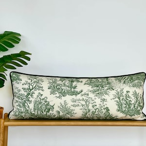 May include: A rectangular decorative pillow with a cream-colored background and a green toile pattern depicting pastoral scenes. The pillow has a dark green border and is placed on a wooden bench.