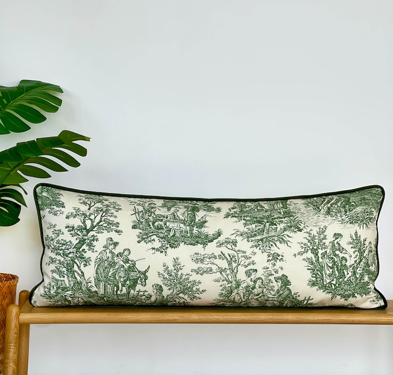 May include: A rectangular decorative pillow with a cream-colored background and a green toile pattern depicting pastoral scenes. The pillow has a dark green border and is placed on a wooden bench.