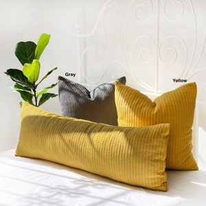 May include: A long yellow bolster pillow with a cable knit texture, placed on a white bed with a white metal headboard. Two smaller square pillows are behind the bolster pillow, one grey and one yellow, both with cable knit texture. The pillows are labelled "Gray" and "Yellow".