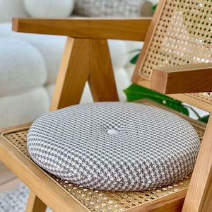 Puede incluir: Un coj&iacute;n de asiento redondo con un estampado de pata de gallo gris y blanco descansa sobre una silla de madera con respaldo de mimbre tejido. El coj&iacute;n tiene un bot&oacute;n en el centro. La silla tiene un acabado de madera clara.