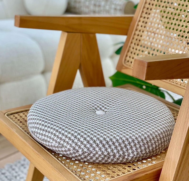Puede incluir: Un coj&iacute;n de asiento redondo con un estampado de pata de gallo gris y blanco descansa sobre una silla de madera con respaldo de mimbre tejido. El coj&iacute;n tiene un bot&oacute;n en el centro. La silla tiene un acabado de madera clara.