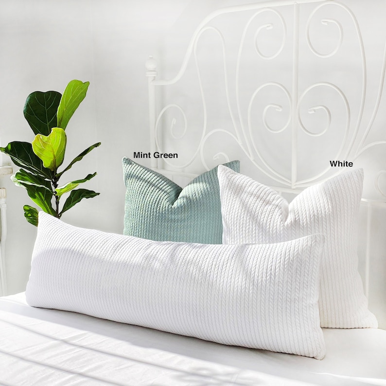 May include: A white, long, rectangular pillow with a cable knit texture on a bed with a white metal headboard. Two square pillows are on the bed, one is mint green and the other is white, both with a cable knit texture.