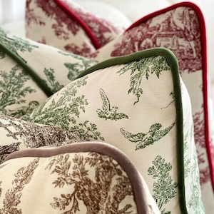 May include: Close-up of decorative throw pillows with a toile pattern. The pillows feature a cream-colored background with green, brown, and red designs of birds, trees, and pastoral scenes. Each pillow has a contrasting colored trim.