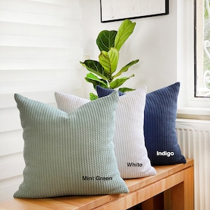 May include: Three decorative throw pillows with a cable knit texture. The pillows are in mint green, white, and indigo colours. The pillows are arranged on a wooden surface.
