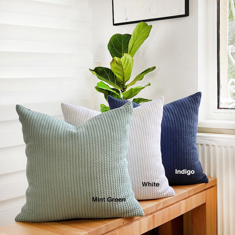 May include: Three decorative throw pillows with a cable knit texture. The pillows are in mint green, white, and indigo colours. The pillows are arranged on a wooden surface.