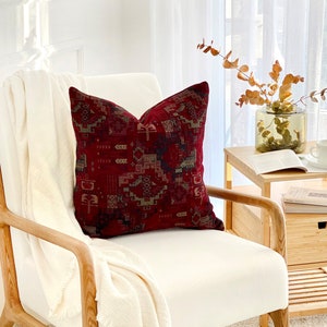 May include: A white armchair with a red and green patterned throw pillow. The chair is made of wood and has a white cushion. The chair is in a room with a white wall and a window with white curtains.