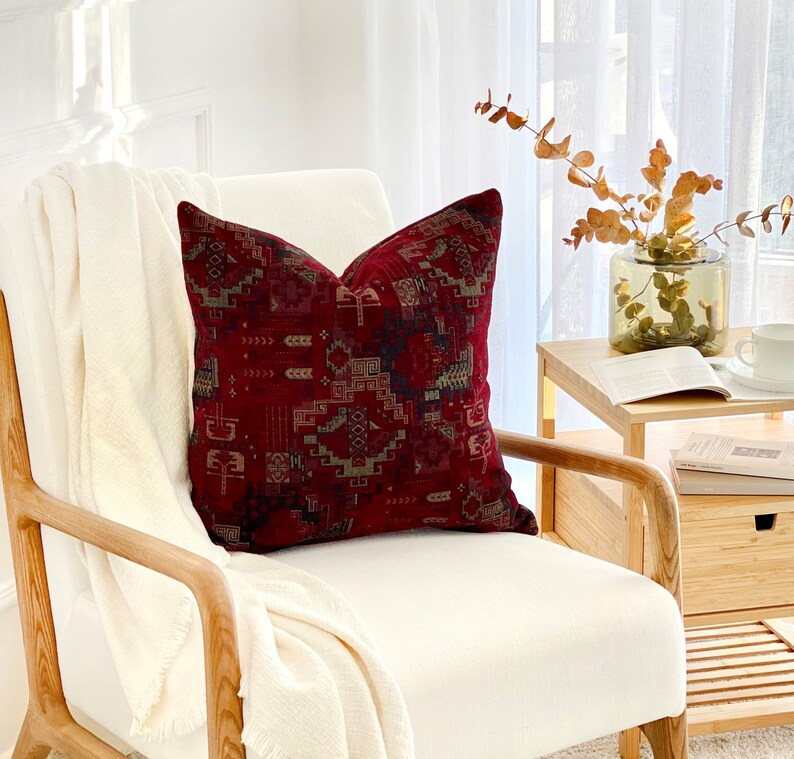 May include: A white armchair with a red and green patterned throw pillow. The chair is made of wood and has a white cushion. The chair is in a room with a white wall and a window with white curtains.