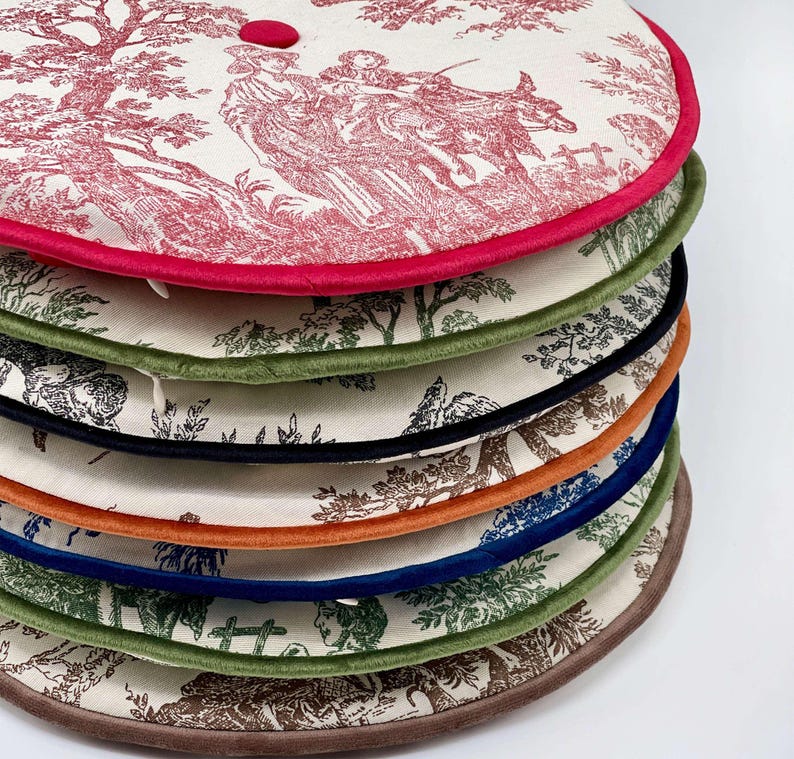May include: A stack of round, decorative cushions with a toile pattern in red and white. Each cushion has a different colored trim, including pink, green, black, orange, blue, and brown. A red button is visible on the top cushion.