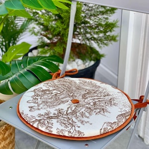 May include: Round chair cushion with a brown toile pattern on a white background, trimmed with orange velvet. The cushion is secured to a gray chair with orange ties. A button is in the center of the cushion.