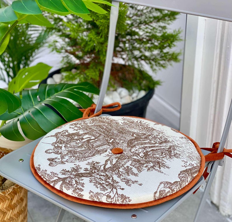 May include: Round chair cushion with a brown toile pattern on a white background, trimmed with orange velvet. The cushion is secured to a gray chair with orange ties. A button is in the center of the cushion.