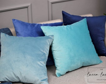 May include: A set of four decorative throw pillows in shades of blue and turquoise. The pillows are made of a soft, velvety fabric and feature a subtle ombre effect. The pillows are arranged on a gray couch with the text "The Lucca Luxury Collection" printed on the couch.