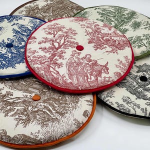 May include: A collection of round cushions with toile patterns in various colors. Each cushion features a different color trim and a button in the center. The cushions display scenes of people and landscapes in shades of blue, red, green, brown, and black.