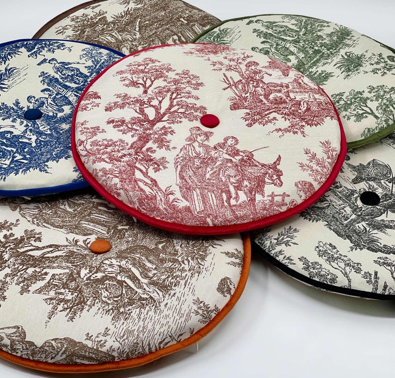 May include: A collection of round cushions with toile patterns in various colors. Each cushion features a different color trim and a button in the center. The cushions display scenes of people and landscapes in shades of blue, red, green, brown, and black.