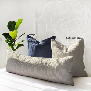 May include: Two decorative throw pillows, one in indigo blue and one in light grey stone, are placed on a long, beige bolster pillow. The pillows are all made of a textured fabric with a cable knit design. The pillows are on a white bed with a white metal headboard.