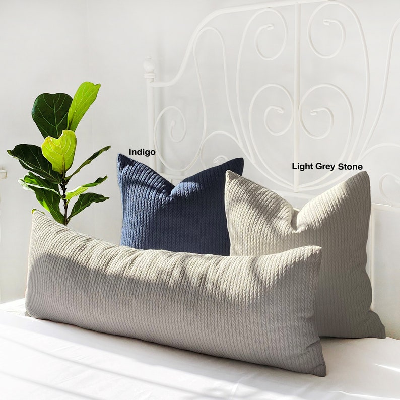 May include: Two decorative throw pillows, one in indigo blue and one in light grey stone, are placed on a long, beige bolster pillow. The pillows are all made of a textured fabric with a cable knit design. The pillows are on a white bed with a white metal headboard.
