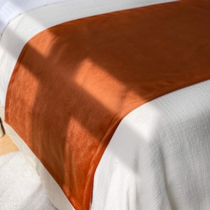 Peut inclure: Un chemin de lit orange br&ucirc;l&eacute; en mati&egrave;re douce et velout&eacute;e. Le chemin de lit est drap&eacute; sur un couvre-lit blanc, cr&eacute;ant un contraste de textures et de couleurs. Un traversin assorti avec une bordure bleue est visible en arri&egrave;re-plan.