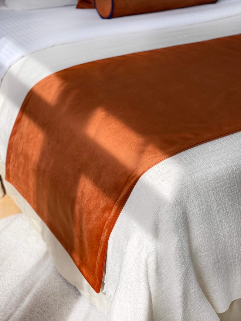 Peut inclure: Un chemin de lit orange br&ucirc;l&eacute; en mati&egrave;re douce et velout&eacute;e. Le chemin de lit est drap&eacute; sur un couvre-lit blanc, cr&eacute;ant un contraste de textures et de couleurs. Un traversin assorti avec une bordure bleue est visible en arri&egrave;re-plan.