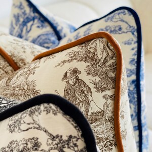 K&ouml;nnte beinhalten: Nahaufnahme von dekorativen Zierkissen mit Toile-de-Jouy-Mustern. Die Kissen zeigen braune und wei&szlig;e pastorale Szenen, blau-wei&szlig;e Blumenmuster und sind mit braunen und marineblauen Paspeln versehen. Die Kissen sind auf einer wei&szlig;en Oberfl&auml;che angeordnet.