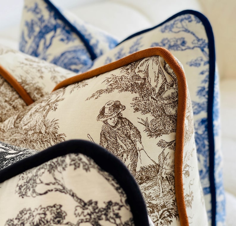 K&ouml;nnte beinhalten: Nahaufnahme von dekorativen Zierkissen mit Toile-de-Jouy-Mustern. Die Kissen zeigen braune und wei&szlig;e pastorale Szenen, blau-wei&szlig;e Blumenmuster und sind mit braunen und marineblauen Paspeln versehen. Die Kissen sind auf einer wei&szlig;en Oberfl&auml;che angeordnet.