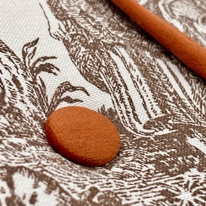 May include: An oval, orange button sits on a cream-colored fabric with a brown toile pattern. A brown, cylindrical object is in the background. The fabric has a detailed illustration of birds and foliage.
