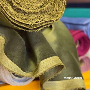 May include: Close-up of moss green fabric rolls. The fabric has a soft, velvety texture and a frayed edge. The text "Moss Green" is visible on the fabric. Other colored fabric rolls are visible in the background.