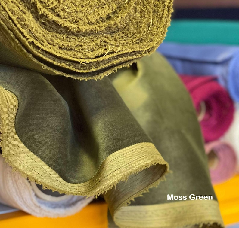 May include: Close-up of moss green fabric rolls. The fabric has a soft, velvety texture and a frayed edge. The text "Moss Green" is visible on the fabric. Other colored fabric rolls are visible in the background.