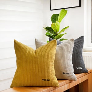 May include: Three decorative throw pillows with a cable knit texture. The pillows are arranged on a wooden bench. The pillows are yellow, stone, and grey. The pillows are labelled "Yellow", "Stone", and "Grey".
