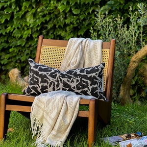 May include: A wooden chair with a woven cane back and seat, a black and white patterned throw pillow, and a cream-colored throw blanket. The chair is sitting on a green grassy lawn.