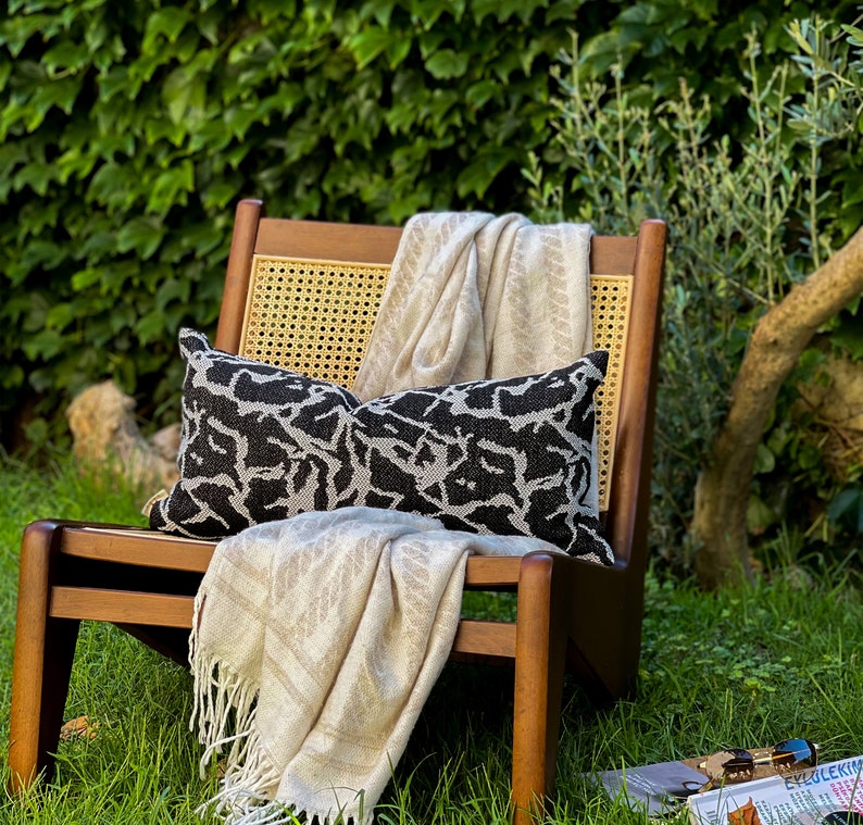 May include: A wooden chair with a woven cane back and seat, a black and white patterned throw pillow, and a cream-colored throw blanket. The chair is sitting on a green grassy lawn.