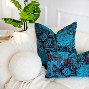 May include: Two teal and brown patterned throw cushions sit on a white sofa. A white round cushion and a white blanket are also present. A green plant is in the background. The cushions are for home decor.