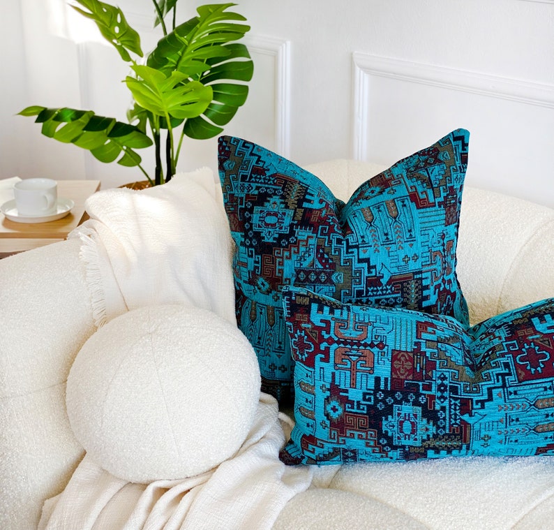 May include: Two teal and brown patterned throw cushions sit on a white sofa. A white round cushion and a white blanket are also present. A green plant is in the background. The cushions are for home decor.