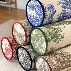 May include: A collection of cylindrical throw pillows with toile patterns in various colors. The pillows feature blue, red, brown, green, and black designs with matching trim. The pillows are arranged on a light-colored rug.