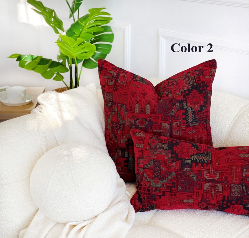 May include: Two decorative throw pillows with a red and black geometric pattern. The pillows are on a white couch with a white throw blanket and a green plant in the background. The text "Color 2" is visible in the top right corner of the image.