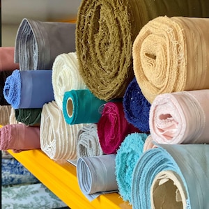 May include: A variety of fabric rolls in different colors, including blue, pink, green, and beige, are stacked on a yellow shelf. The rolls are tightly wound, showcasing the texture and color variations of the textiles. The image highlights the diverse range of materials available.