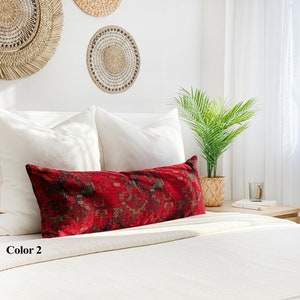 May include: A long, rectangular red cushion with a detailed, geometric pattern in black and gold tones rests on a white bed. The cushion is placed against white pillows, with woven wall decor and a potted plant in the background.