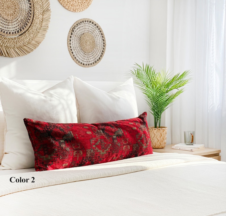 May include: A long, rectangular red cushion with a detailed, geometric pattern in black and gold tones rests on a white bed. The cushion is placed against white pillows, with woven wall decor and a potted plant in the background.