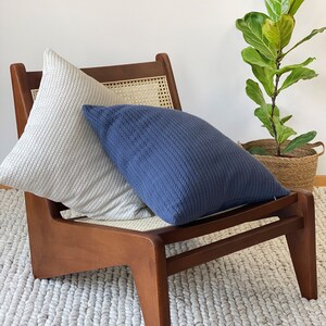 May include: A wooden chair with a woven cane seat and back, featuring two cushions. One cushion is light grey and the other is dark blue. The chair is sitting on a white fluffy rug.